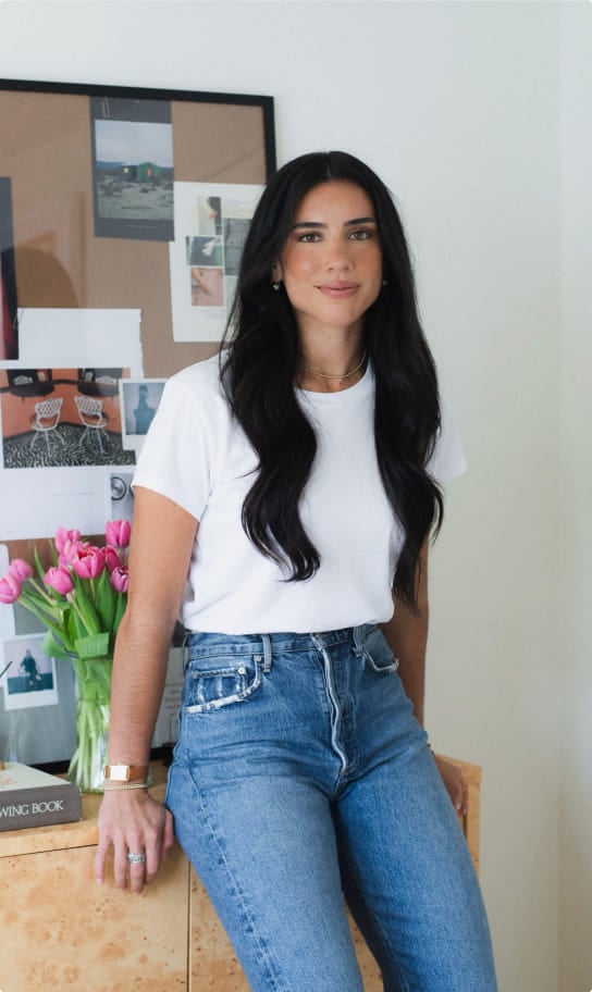 Ceci in a white t-shirt and vintage-wash jeans, leaning against a dresser